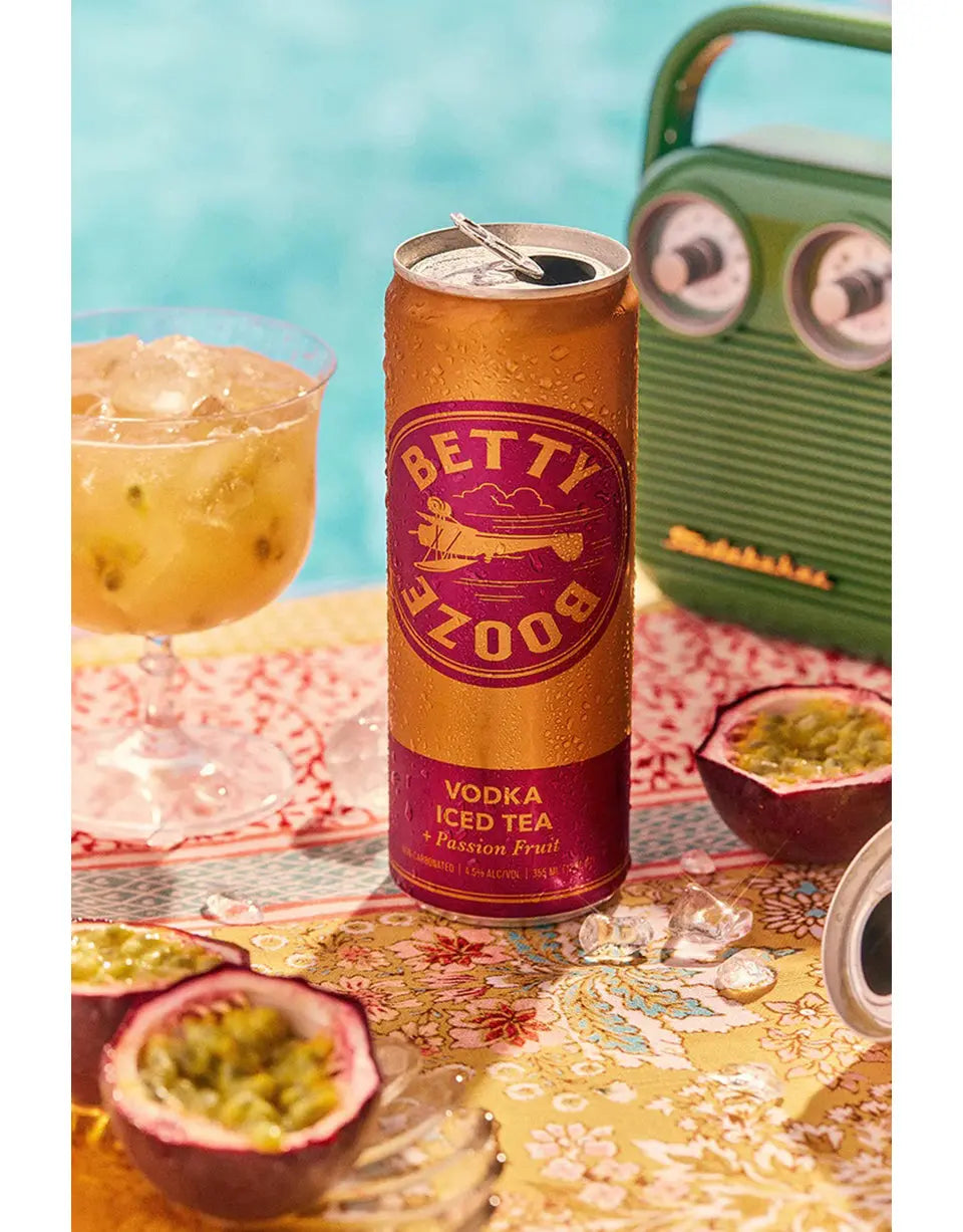 Can of Betty Booze Vodka Iced Tea with Passion Fruit on a table with a cocktail and fruit.