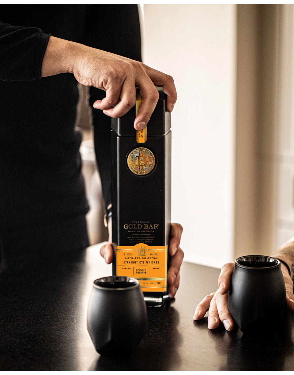 Person holding a bottle labeled 'Gold Bar' with two black cups on a table.