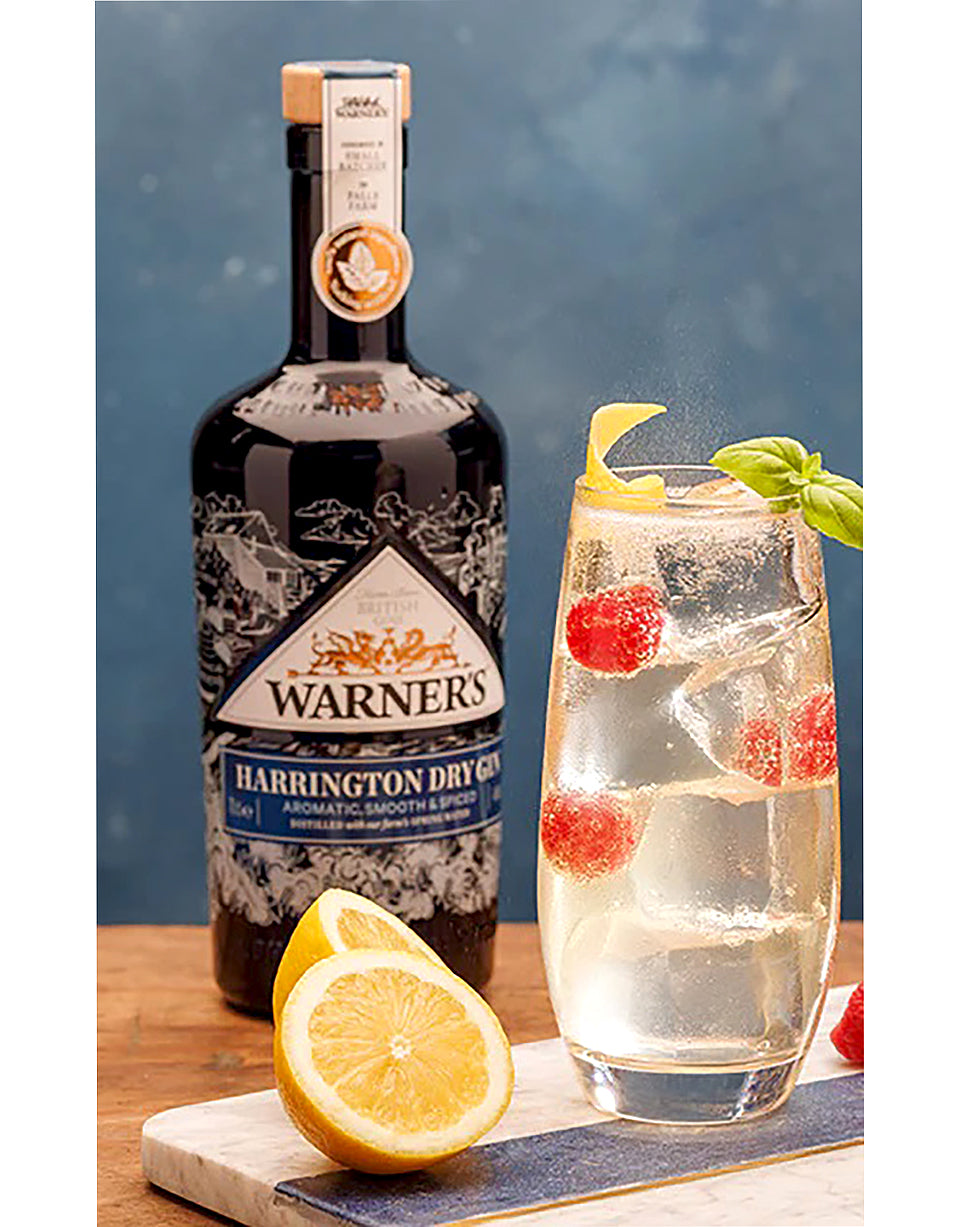Warner's Harrington Dry Gin bottle with a cocktail garnished with lemon and berries on a wooden surface.