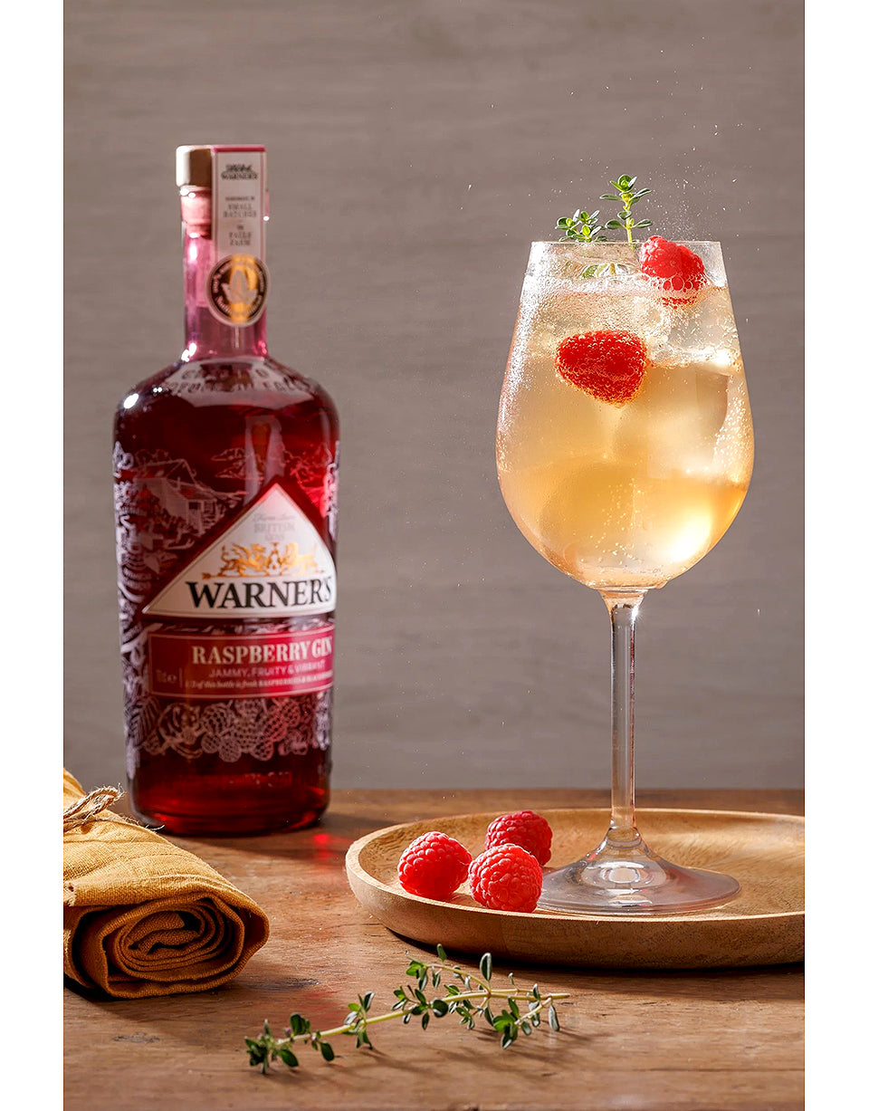 Warner's Raspberry Liqueur bottle with a glass of cocktail garnished with raspberries on a wooden surface.